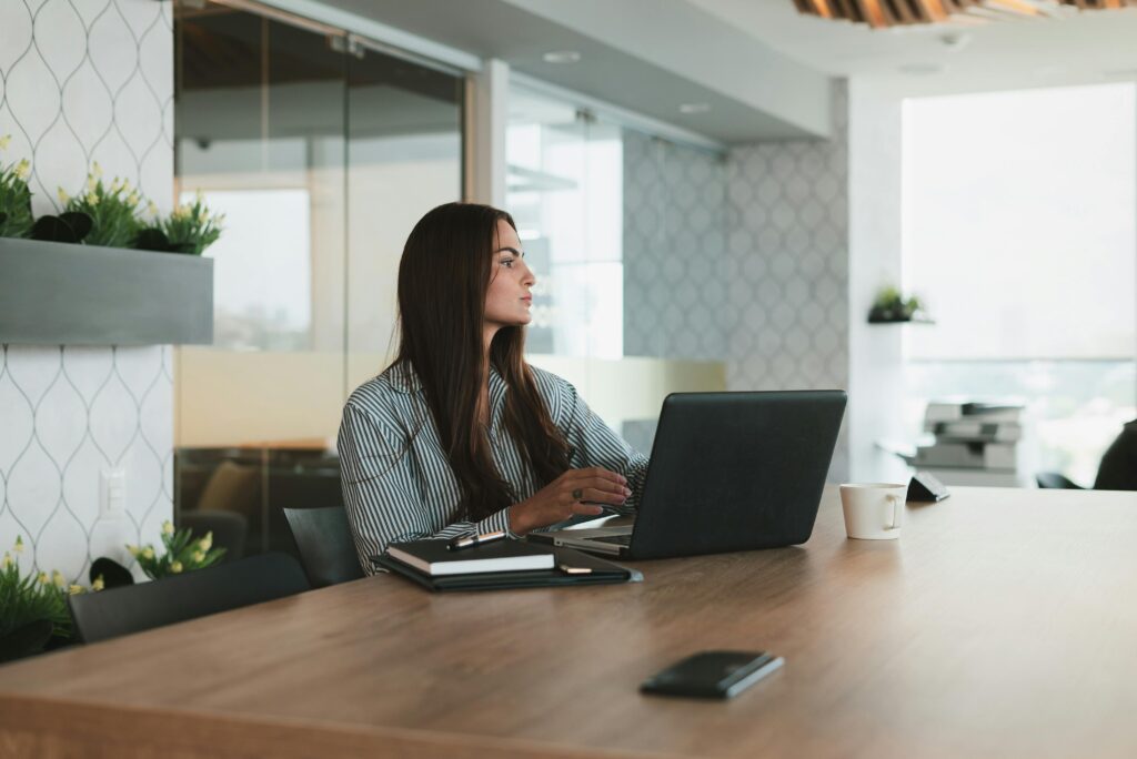 Businesswoman at a desk with a laptop, looking contemplatively out the window.